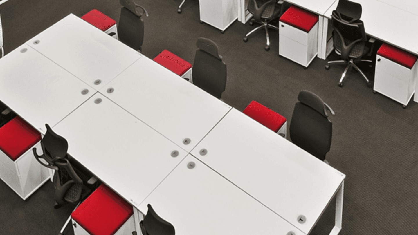 A modern office space with white desks arranged in a rectangular formation. Black office chairs and red cushioned stools are placed around the desks. The floor is carpeted in dark grey. The setting appears organized and minimalistic.