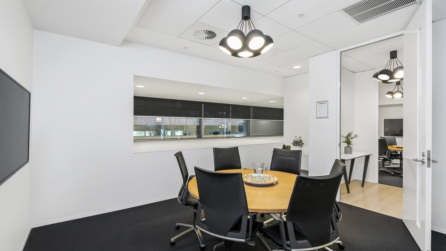 Modern conference room with a round wooden table surrounded by black chairs. Overhead, there are stylish, multi-bulb pendant lights. A flat-screen TV and large window are on the white walls. Door leading to another office space is open.