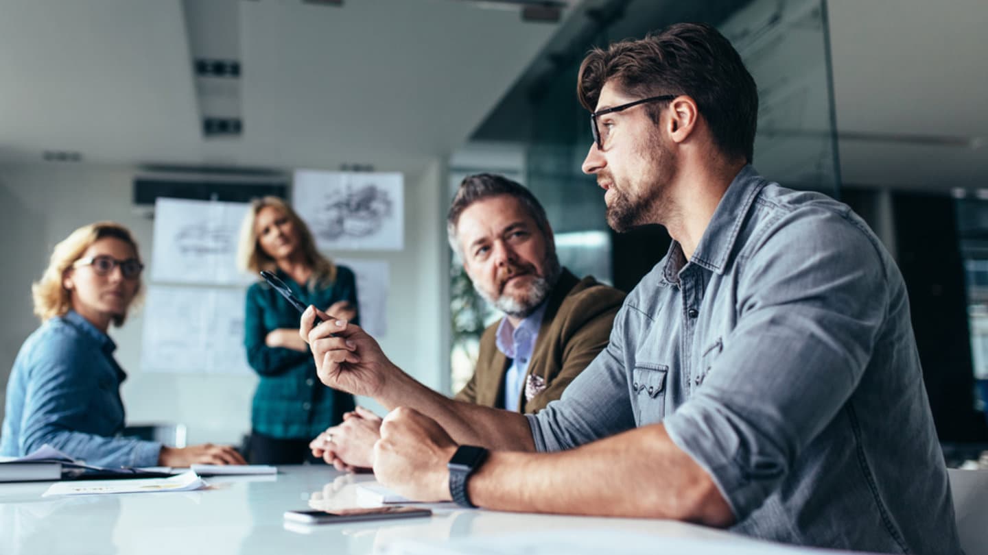 A group of four people sit around a conference table in an office. One person is speaking while others listen attentively. Papers and a notebook are on the table, and diagrams are displayed on the glass wall in the background.