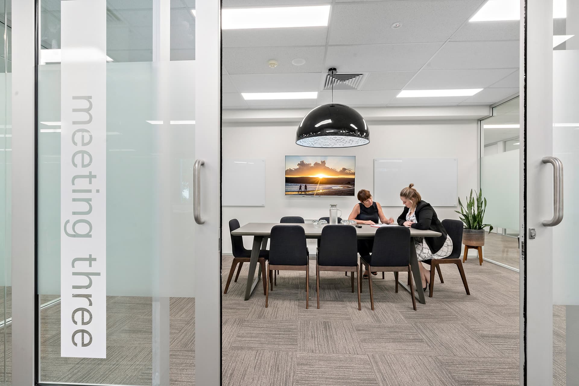 View through the glass door of a modern office meeting room where two women are seated at a table, reviewing documents. The room features white walls, a decorative photo on a screen, and a large pendant light. The door glass has the words "Meeting three".