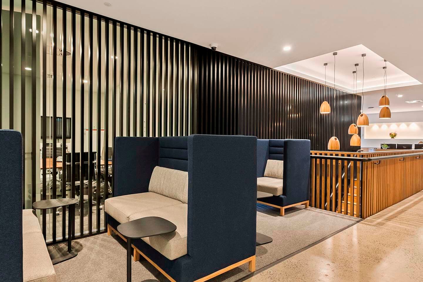 Modern office lounge area with two high-backed booths and small round tables, separated by vertical black slats. In the background, a meeting room with glass walls and pendant lights above a staircase. Neutral tones dominate the decor.