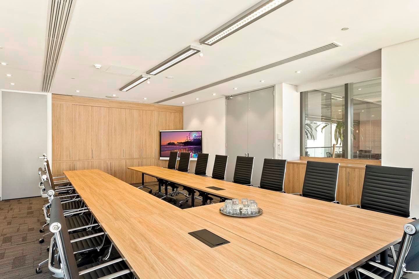 A modern conference room with a large wooden table surrounded by black chairs. A flat-screen TV displays an image of a bridge. The room features light wood paneling and has a bright atmosphere with overhead lighting.