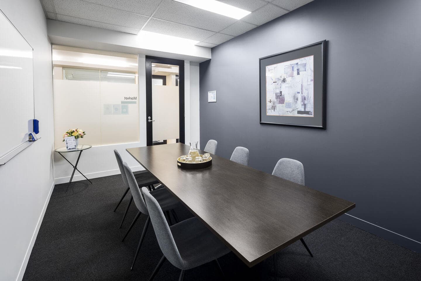A modern conference room with a long dark wooden table and six gray chairs. Theres a whiteboard on the left wall, a framed abstract painting on the right wall, and a small table with flowers near the door. The room has gray carpeting.