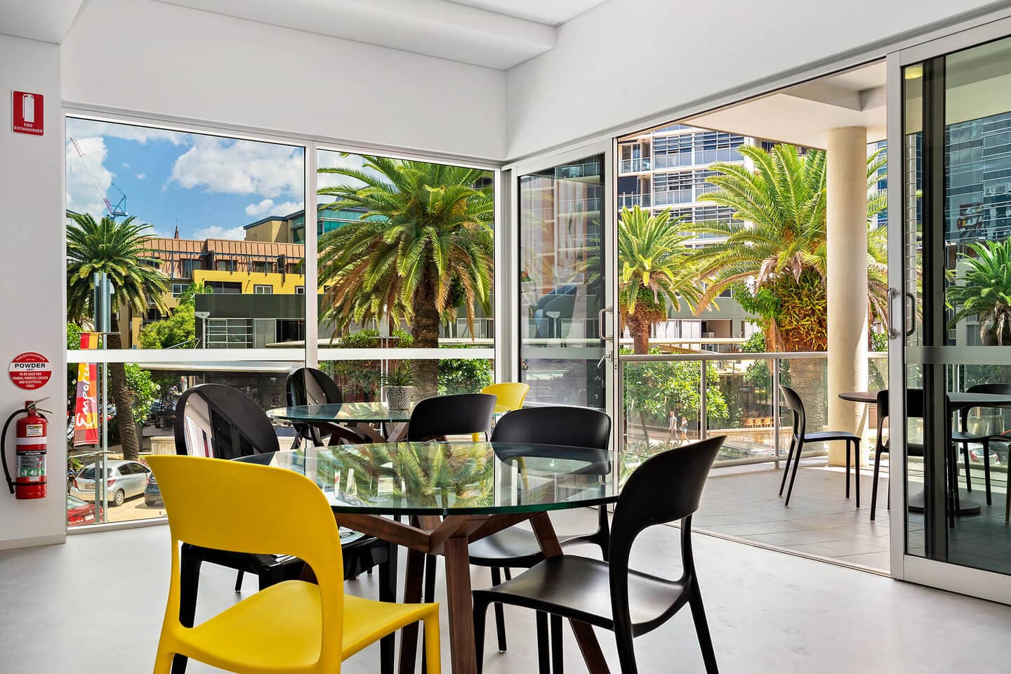 A bright indoor seating area with glass tables and modern black and yellow chairs. Large windows and sliding doors reveal a balcony with palm trees and urban buildings outside. A fire extinguisher is mounted on the white wall.