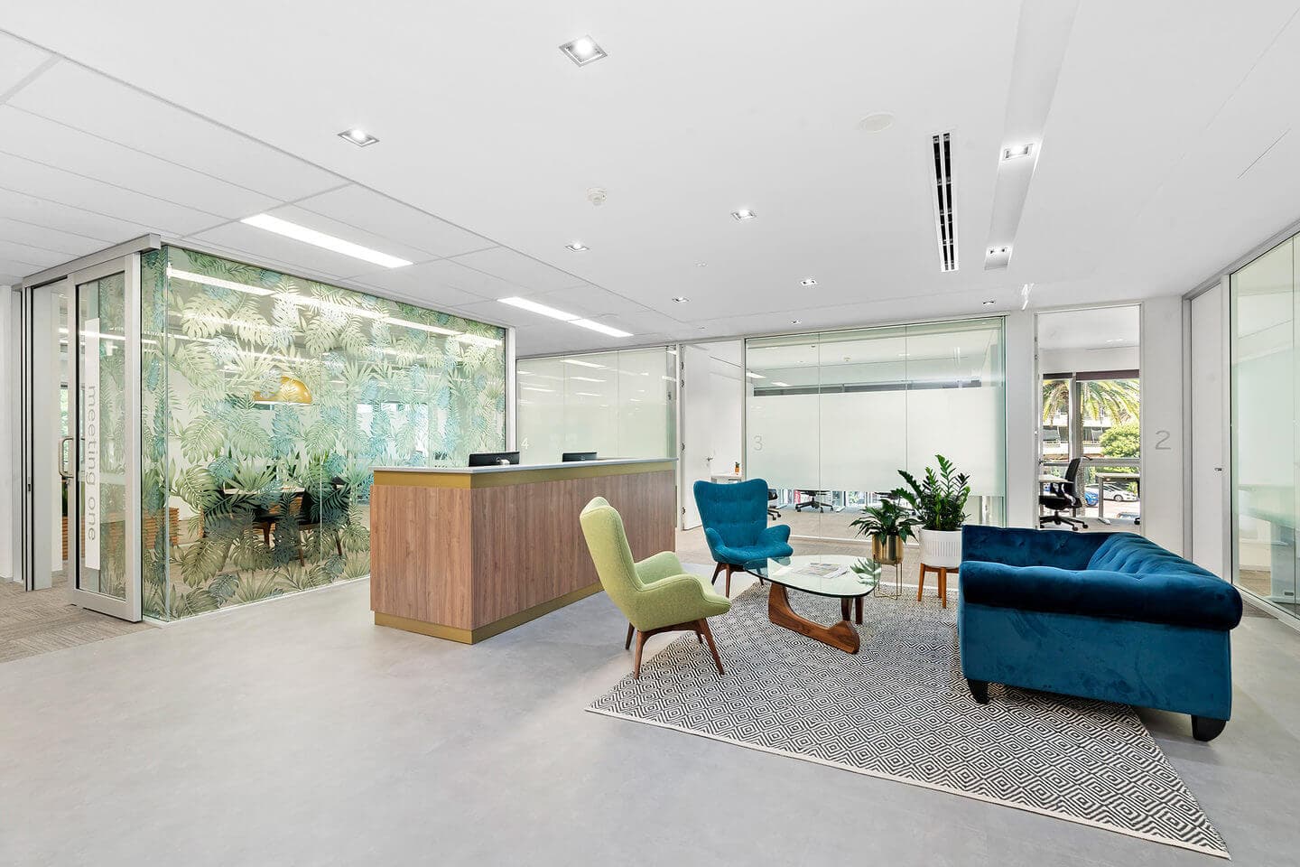 Modern office reception area with a wooden desk on the left side. Includes a green and a blue armchair, a blue sofa, and a glass table on a patterned rug. Glass walls display tropical-themed designs on the left and offer a view of another office area.