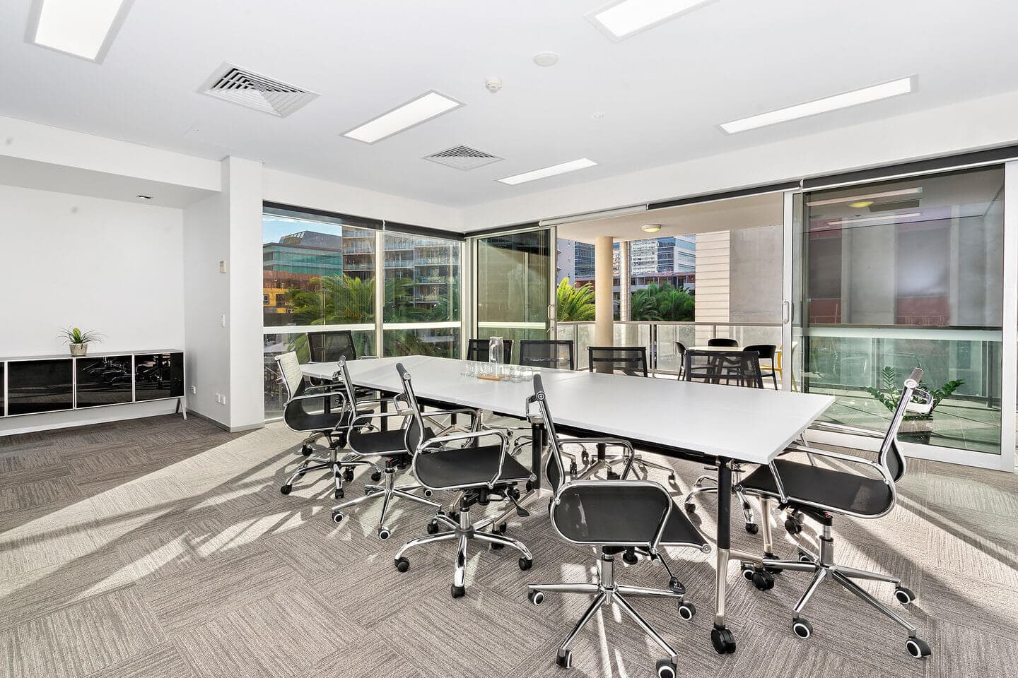 Modern conference room with a long white table surrounded by black swivel chairs. Large windows open to a view of nearby buildings, allowing natural light to fill the room. Neutral-toned carpet and minimal decor create an open, airy atmosphere.