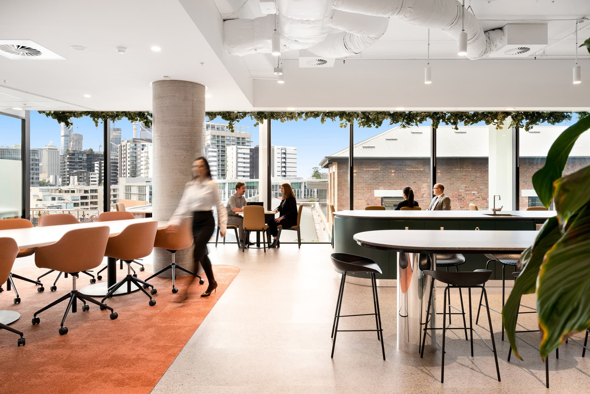 Modern office space with round tables and chairs, a high counter with stools, and large windows overlooking a cityscape. A person walks through the area, while others sit and work or converse near the windows. Greenery decorates the ceiling edge.