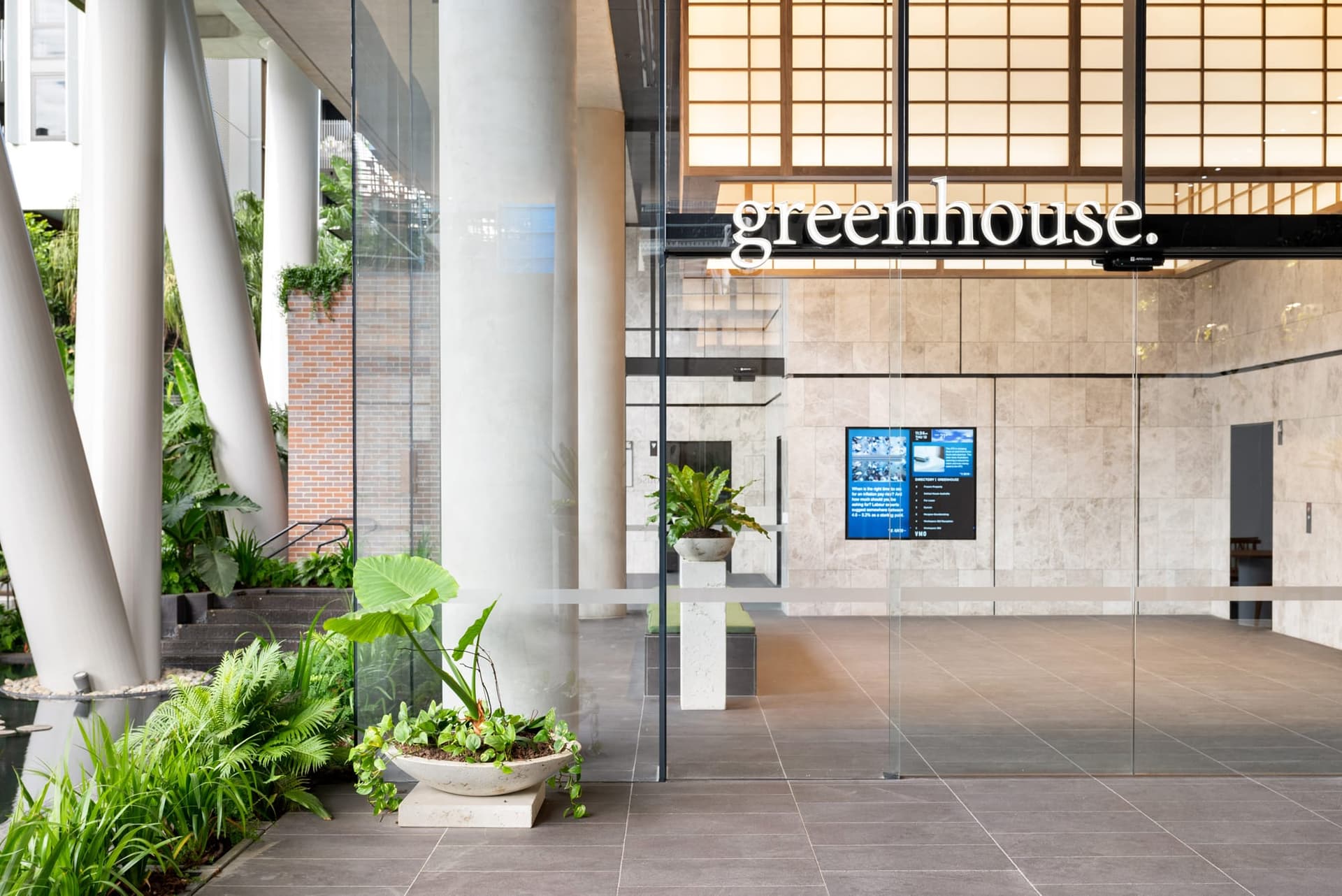 Entrance to a modern building with large glass windows and greenhouse text on the facade. Inside, there are plants, a screen displaying information, and stone walls. Outside, more greenery is visible.