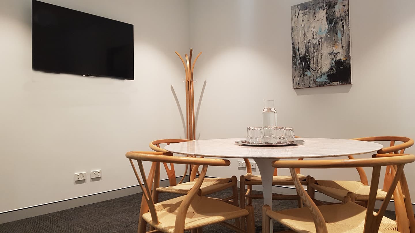 A minimalist meeting room with a round white table surrounded by four wooden chairs. A flat-screen TV is mounted on the wall above a coat rack. A large abstract painting adorns the adjacent wall. Natural lighting brightens the space.