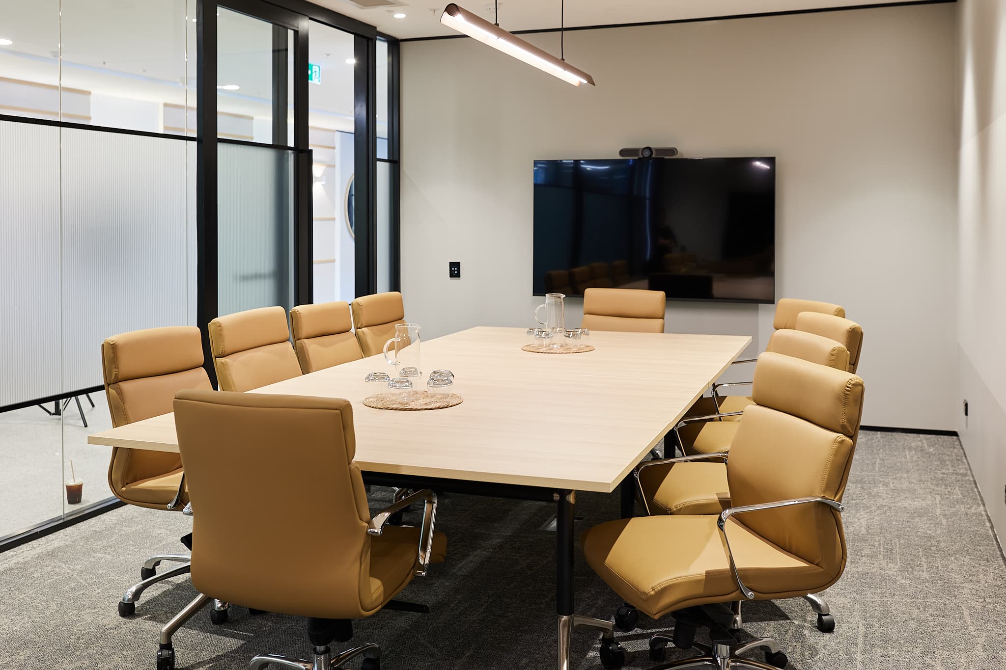 A modern conference room with a rectangular wooden table surrounded by eight tan leather chairs. A large flat-screen TV is mounted on the wall. The room has glass walls and neutral decor, with a ceiling light fixture above the table.