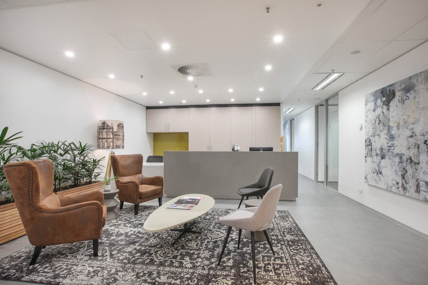 Modern office lobby with a reception desk and stylish seating area. Includes two brown leather armchairs, a white oval coffee table, a gray rug with intricate patterns, and a black chair. A large abstract painting and potted plants decorate the space.