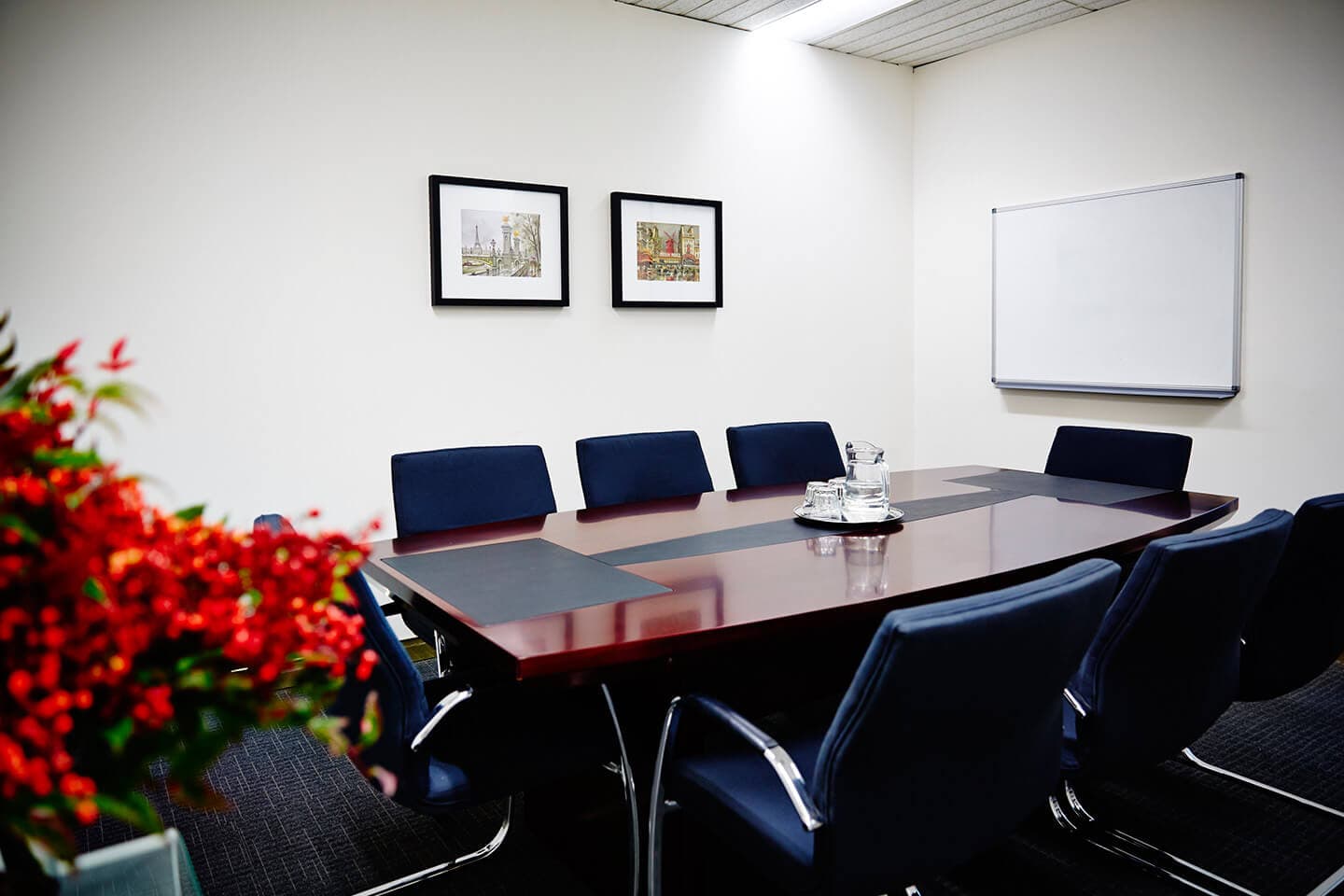 A meeting room with a long wooden table, lined with eight navy blue chairs. A whiteboard is mounted on the right wall, and two framed pictures are on the back wall. Red flowers are in the foreground on the left.