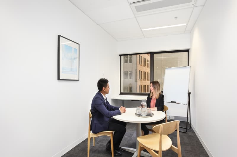Two people sit across from each other at a small round table in a modern office. One person wears a suit and the other wears business casual attire. The room is bright with a window, a blank flip chart, and a framed picture on the wall.