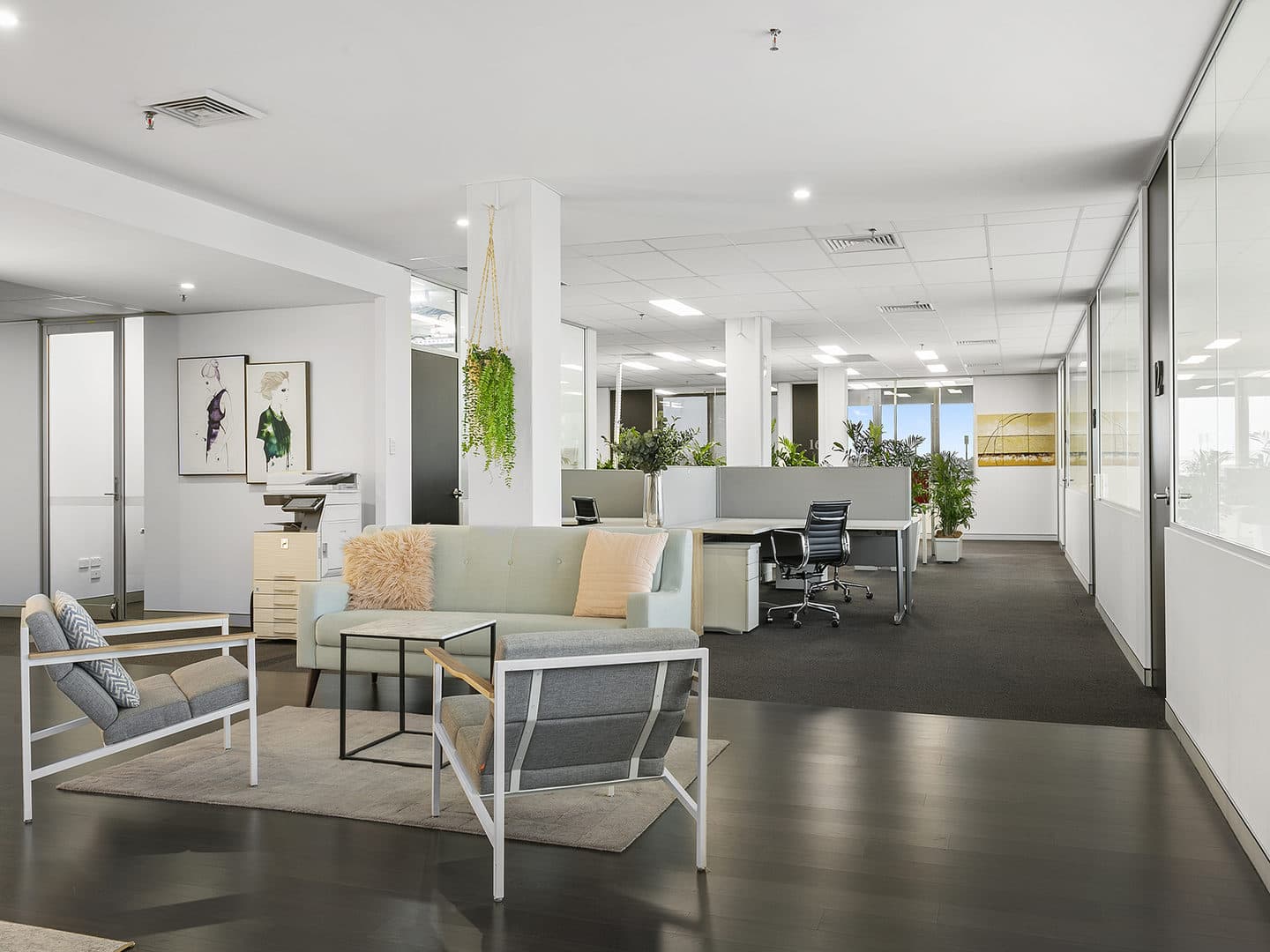 Modern open-plan office with a light green sofa, two gray chairs, and a rug in a seating area. Potted plants and artwork adorn the space. Desks with chairs are in the background, and large windows allow natural light to enter.