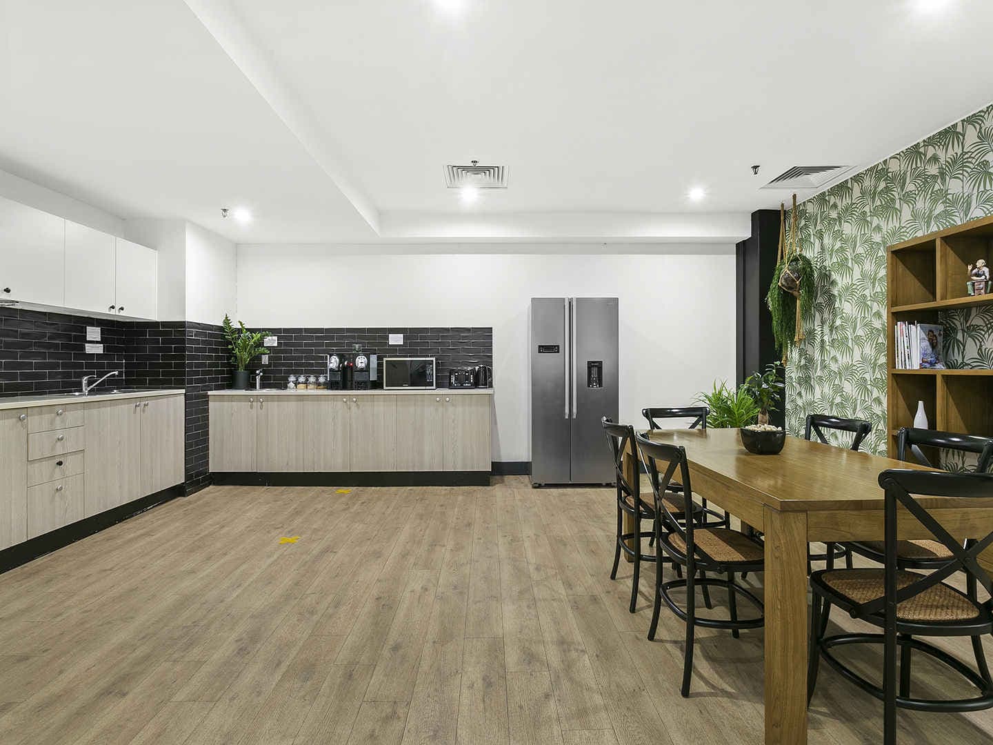 A spacious kitchen and dining area with wooden flooring. It features white and black cabinets, a stainless steel refrigerator, a microwave, and a dining table with six chairs. One wall has tropical leaf-patterned wallpaper and a wooden shelf.