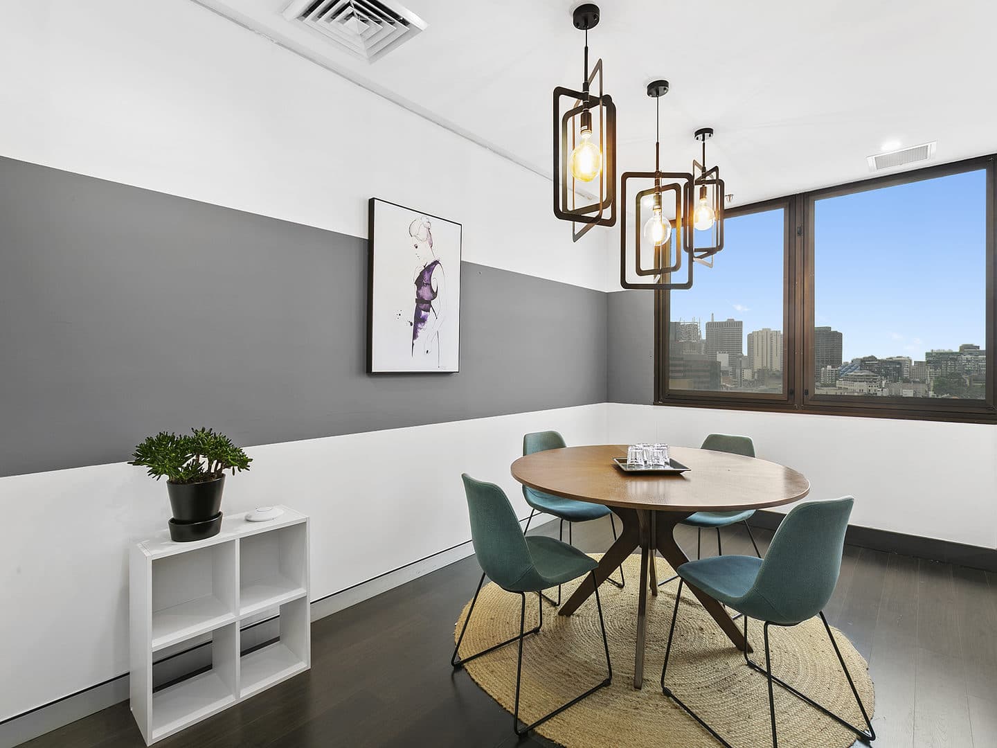 A modern dining room with a round wooden table, four teal chairs, and a geometric chandelier. A potted plant sits on a small white shelf. The walls are gray and white, with a cityscape visible through a large window. A painting decorates the wall.