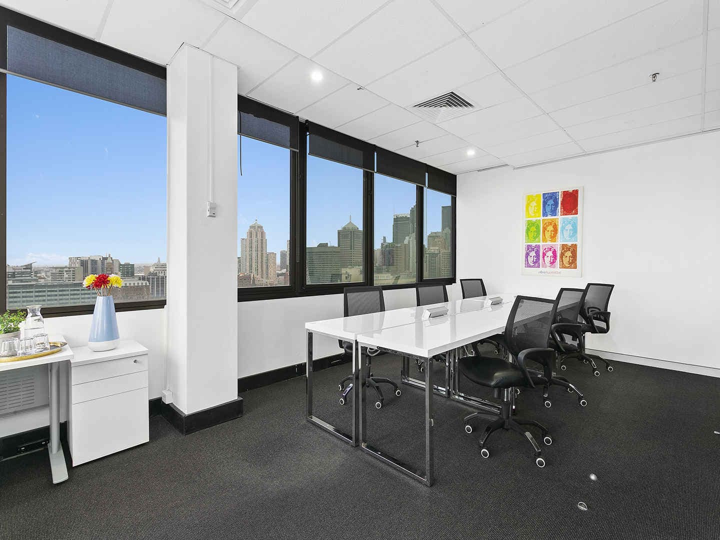 A modern office space with a white desk and six black swivel chairs. Large windows offer a cityscape view. A colorful artwork is displayed on the wall, and a side table holds a vase with flowers. The room is well-lit with overhead lights.