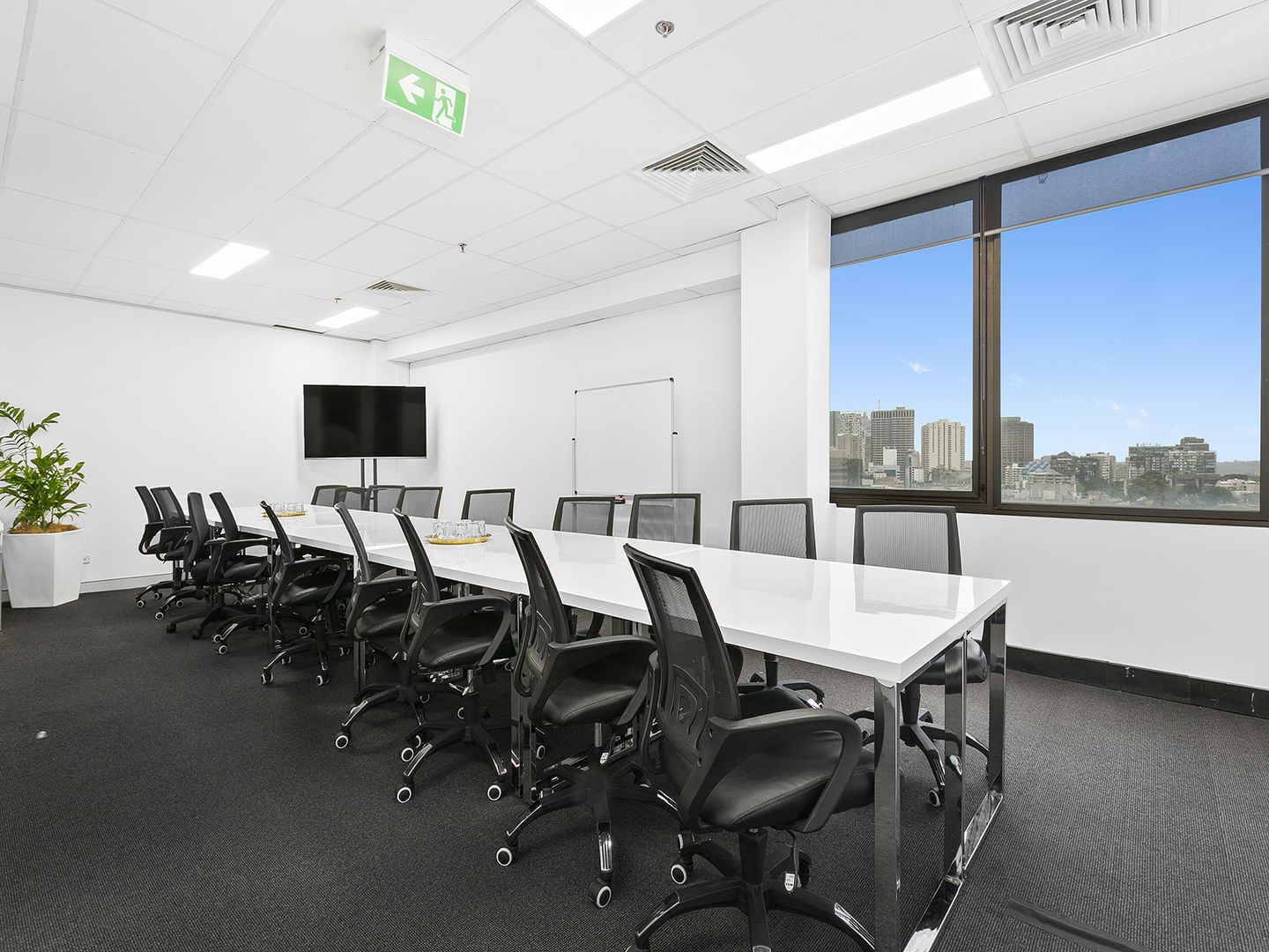 A modern conference room with a long white table surrounded by black office chairs. A large window on the right shows a cityscape view. A flat-screen TV is mounted on the wall, and a potted plant is in the corner.