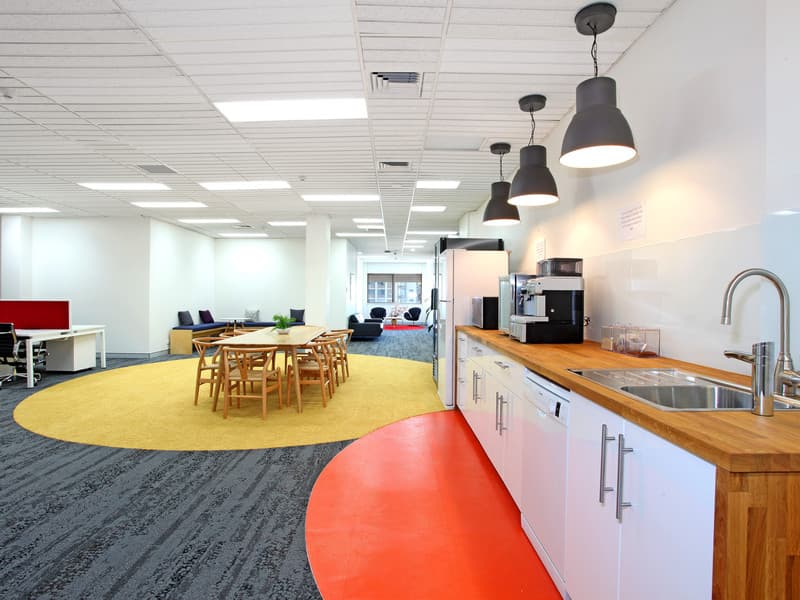 Modern office space with a kitchenette featuring a wooden countertop, coffee machine, and sink. A round table with wooden chairs sits on a yellow rug. Open workstations and lounge chairs are visible in the background, with ample natural light.