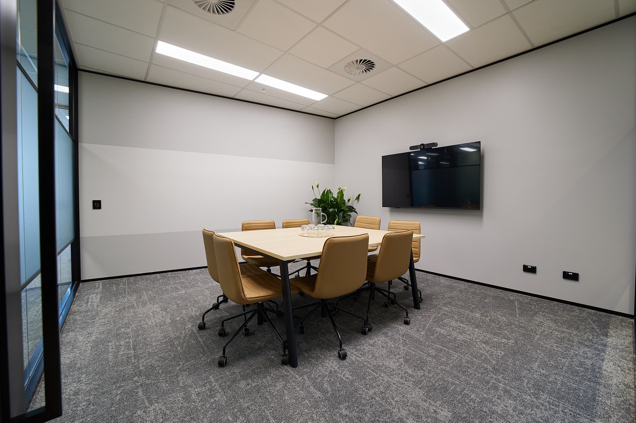 A modern conference room with a rectangular table surrounded by five tan chairs on wheels. A large flat-screen TV is mounted on the wall, and a potted plant is placed in the center of the table. The room has a carpeted floor and white walls.