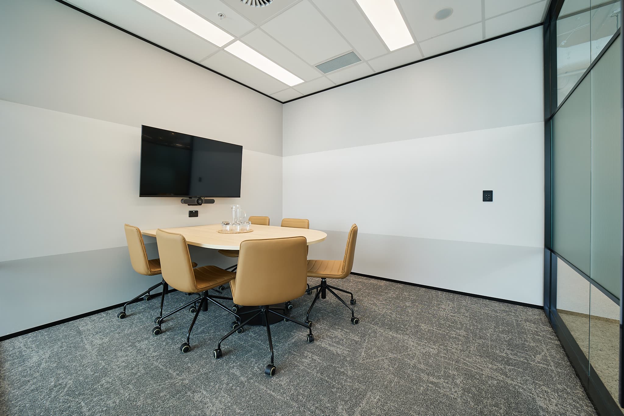 A small, modern conference room with a round table and five tan chairs on wheels. A flat-screen TV is mounted on the white and gray walls. The floor is carpeted, and there is a glass partition on one side.