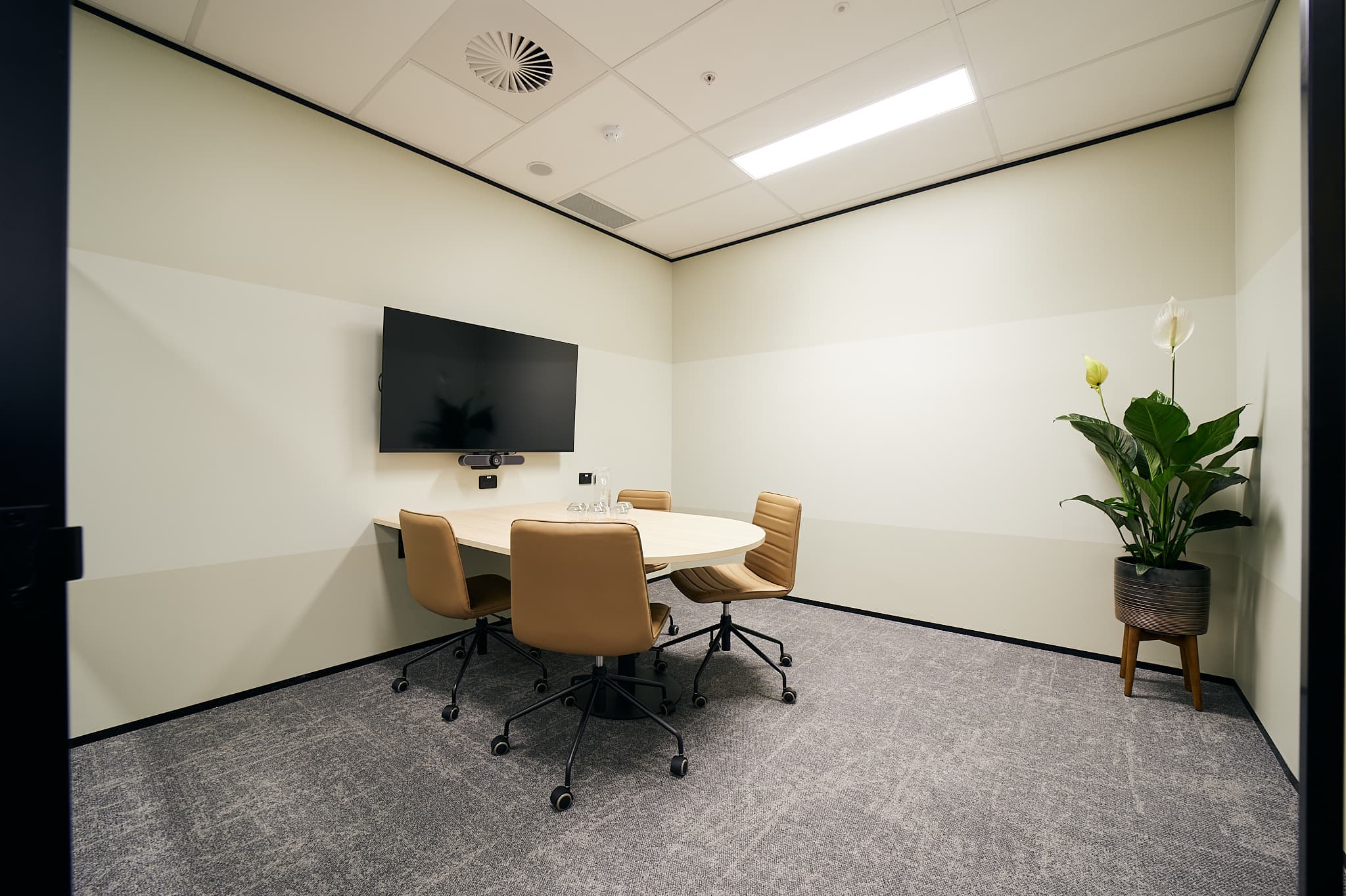 A small conference room with a round table and four beige chairs. A flat-screen TV is mounted on the wall. A potted plant is in the corner. The room has a carpeted floor and neutral-colored walls.
