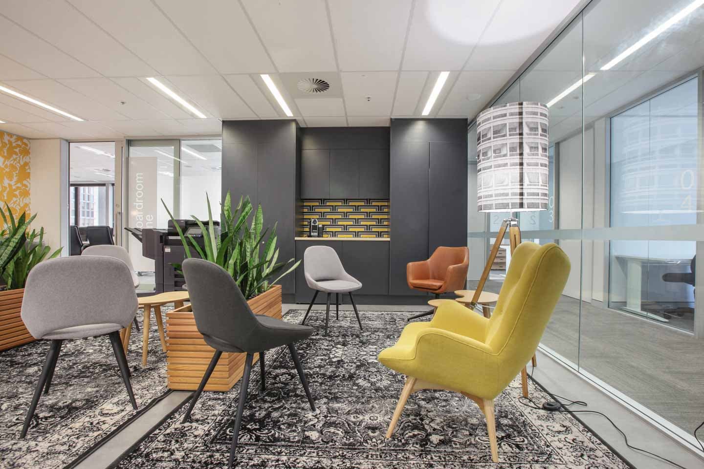 Modern office lounge with various colorful chairs, potted plants, and a patterned rug. Glass walls on the right offer a view into another room. A unique floor lamp stands next to the yellow chair. Bright, inviting atmosphere.