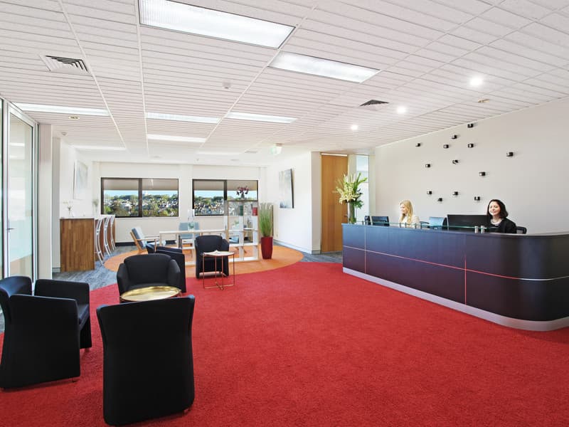 Modern office reception with two women at a curved desk on the right. Red carpet covers the floor, with black chairs and small tables on the left. The back area features a seating area with a round rug, multiple chairs, and windows with outdoor views.