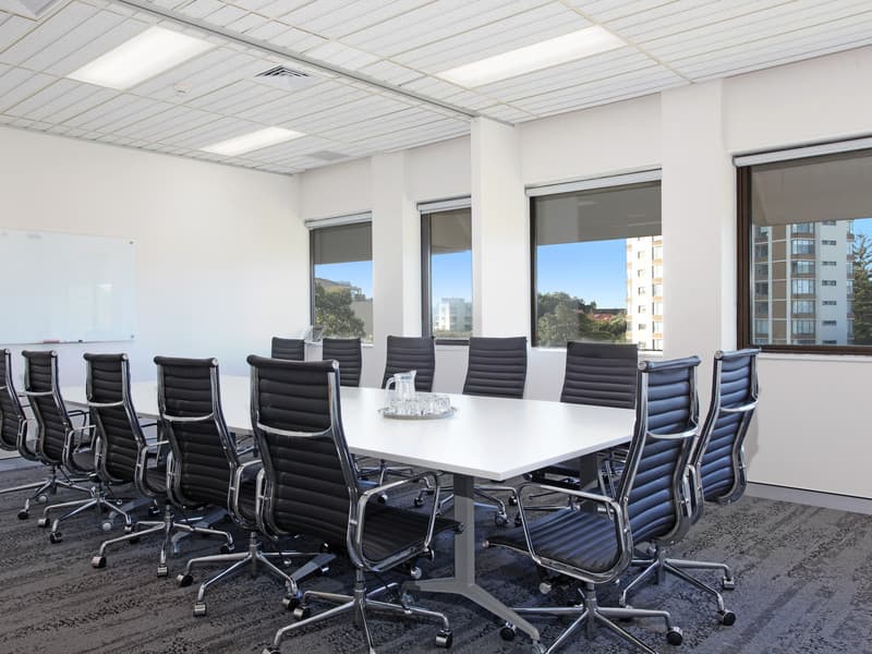 A modern conference room with a long white table and several black chairs arranged around it. Large windows line one side of the room, allowing natural light to enter. A glass water pitcher and glasses are on the table. A whiteboard is on the wall.