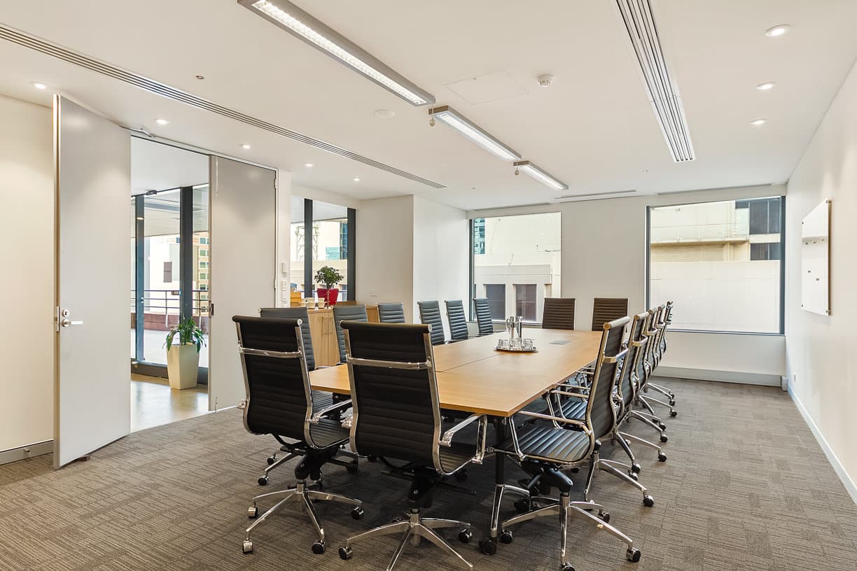 A modern conference room with a long wooden table surrounded by twelve black leather office chairs. Two large windows provide natural light, and a glass door opens to a balcony. The room has recessed lighting and a carpeted floor.