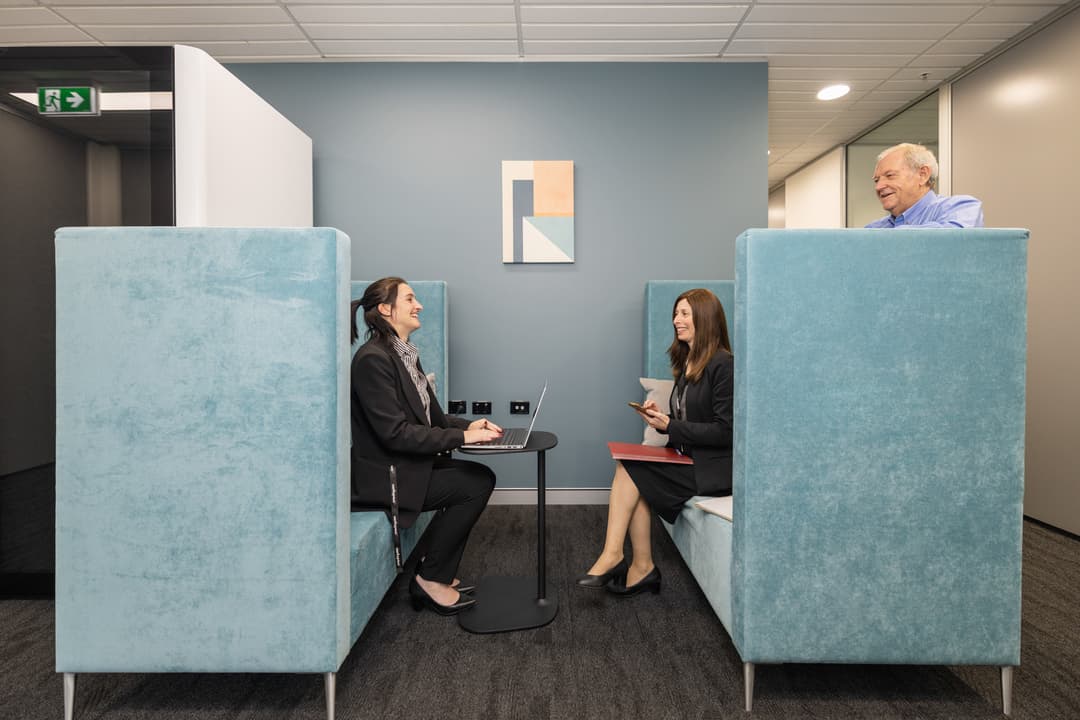 Three people in business attire are having a meeting in a modern office lounge area with blue booth seating, a small black table, and abstract wall art. One person stands, while two sit and use a laptop and notepad.