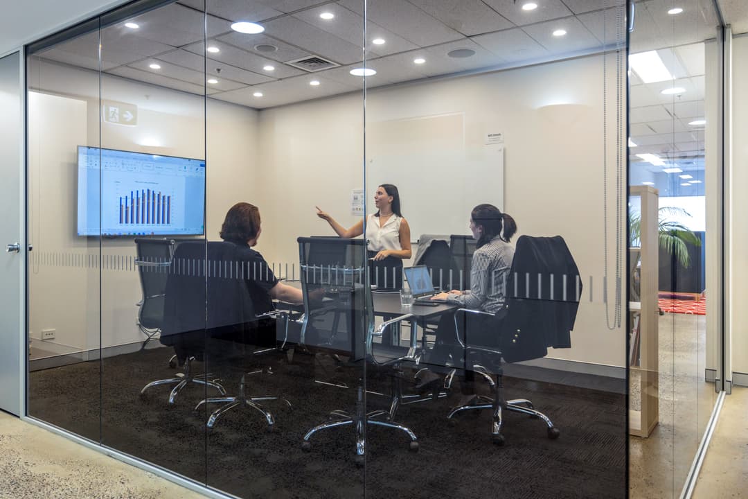 Three people sit at a conference table in a glass-walled meeting room while a fourth person stands and presents a bar graph on a screen.
