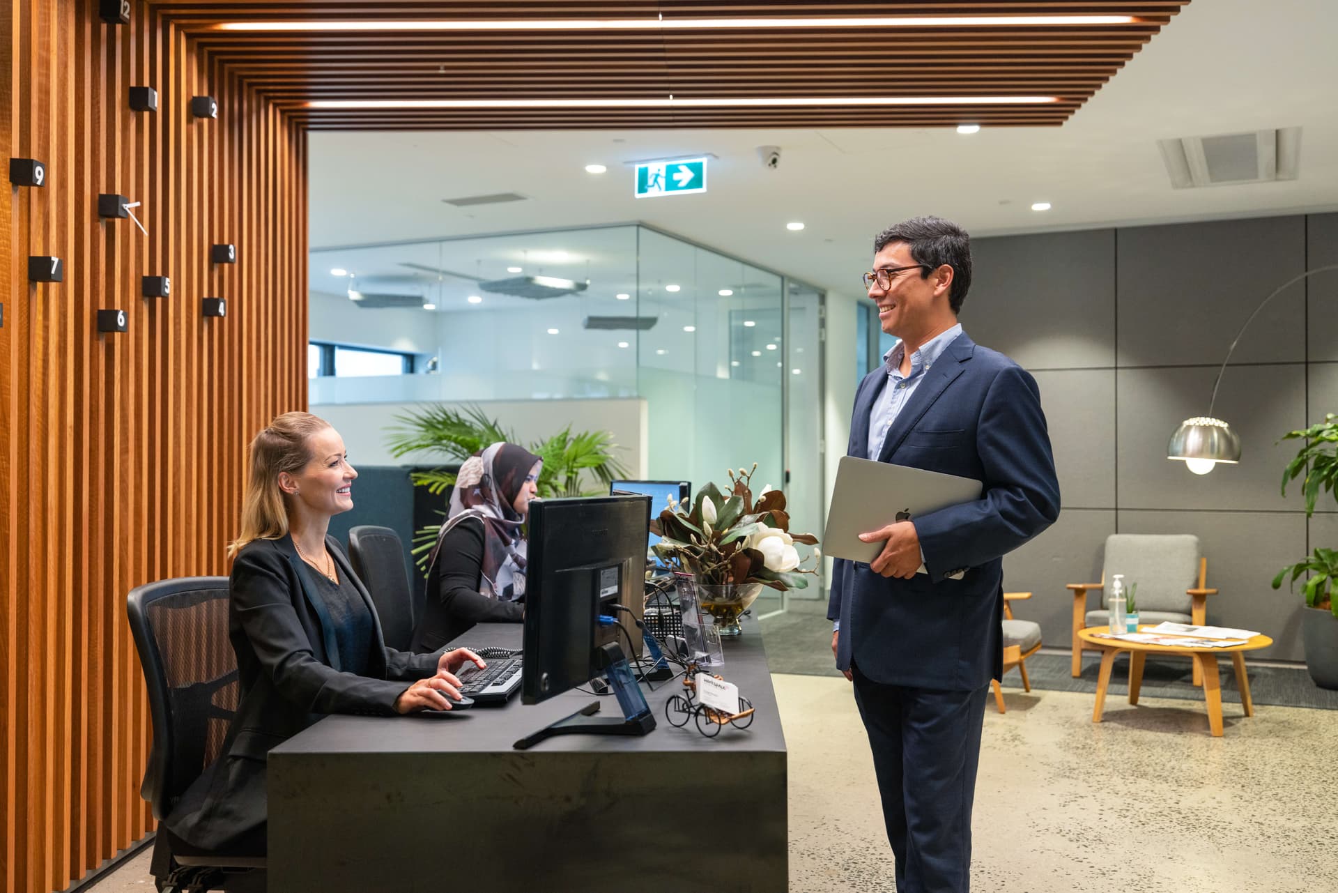 A man in a suit holding a laptop smiles and talks to a receptionist at a modern office front desk, while another woman works at a nearby computer. The office features glass walls and wooden accents.