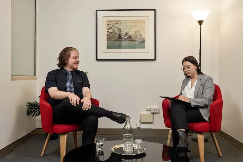 A man and a woman sit on red chairs in a modern office. The man is smiling and relaxed, while the woman is focused, holding a clipboard. A lamp and framed artwork are on the wall behind them. A table with water glasses is in the foreground.