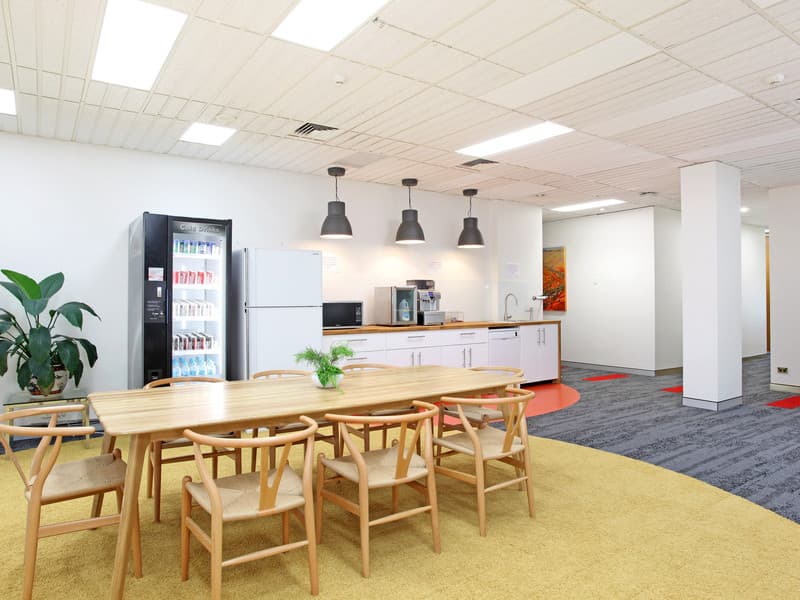 A modern office break room featuring a wooden table with chairs on a circular yellow rug. A vending machine and refrigerator stand against a wall. White cabinets with a coffee machine are under three black pendant lights. A plant and artwork decorate the space.
