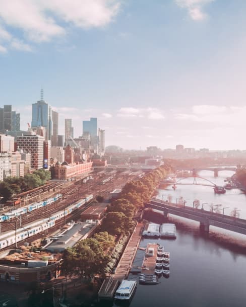 Aerial view of a cityscape featuring modern skyscrapers, a river with docked boats, and multiple railway tracks. Bridges span the river, trees line the waterfront, and the sky is clear with scattered clouds.