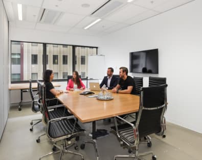 Four people are seated around a large wooden conference table in a modern office. They are engaged in a meeting, with a laptop open on the table. The room has large windows, a flat-screen TV, a whiteboard, and ergonomic chairs.