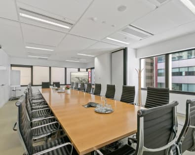 A long, wooden conference table surrounded by black leather office chairs in a modern meeting room with large windows. The room features natural lighting with a view of urban buildings outside. A whiteboard and coat rack are visible in the background.