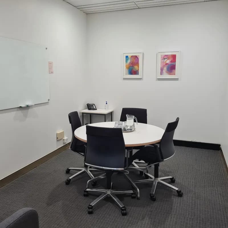 A small conference room with four chairs around a circular table. A pitcher with glasses is on the table. A whiteboard is mounted on the wall to the left. Two abstract art pieces hang on the white wall above a small corner table.
