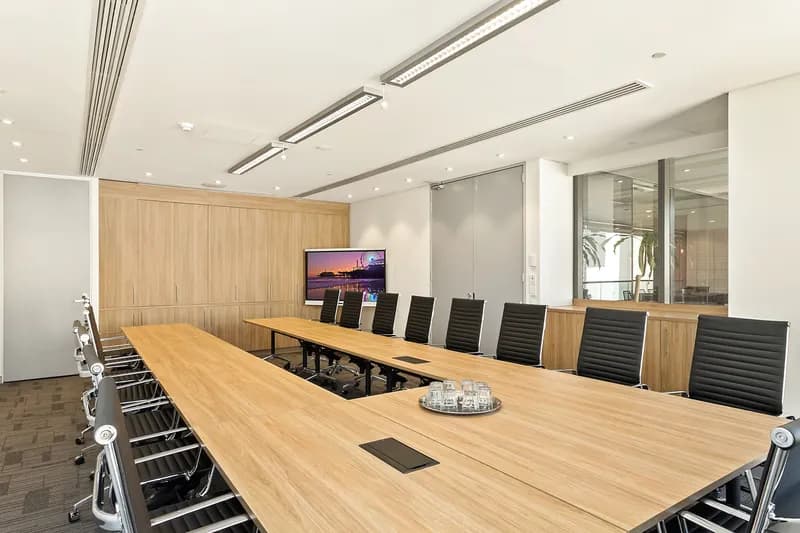 A modern conference room with a long wooden table surrounded by black chairs. A large TV screen at the end of the room displays a cityscape image. The room has beige walls, recessed lighting, and a glass partition on one side.