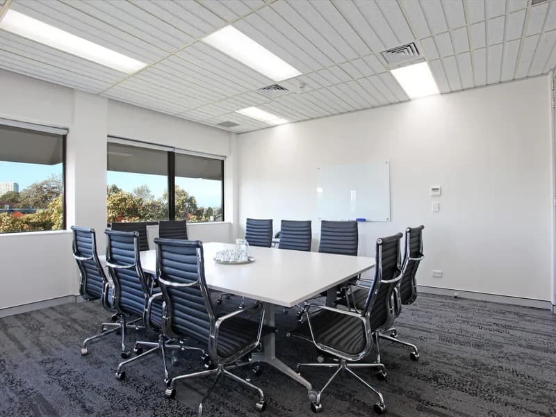 A modern conference room with a long rectangular table surrounded by ten black swivel chairs. Two large windows let in natural light. A whiteboard is mounted on one wall, and a glass pitcher with water and glasses sits on the table.