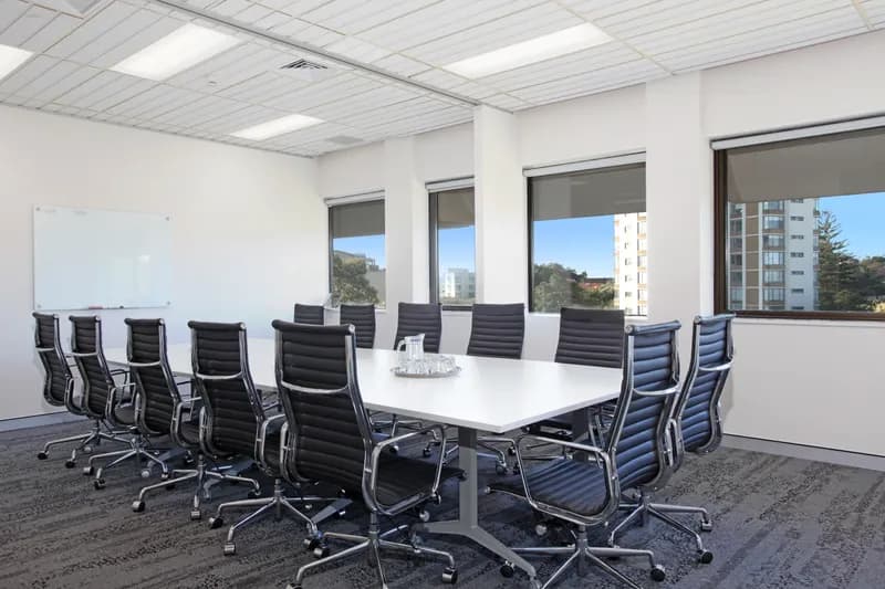 A modern conference room with a long white table surrounded by twelve black leather office chairs. There are large windows showing a view of nearby buildings, and a blank whiteboard on the wall. A tray with glasses sits on the table.