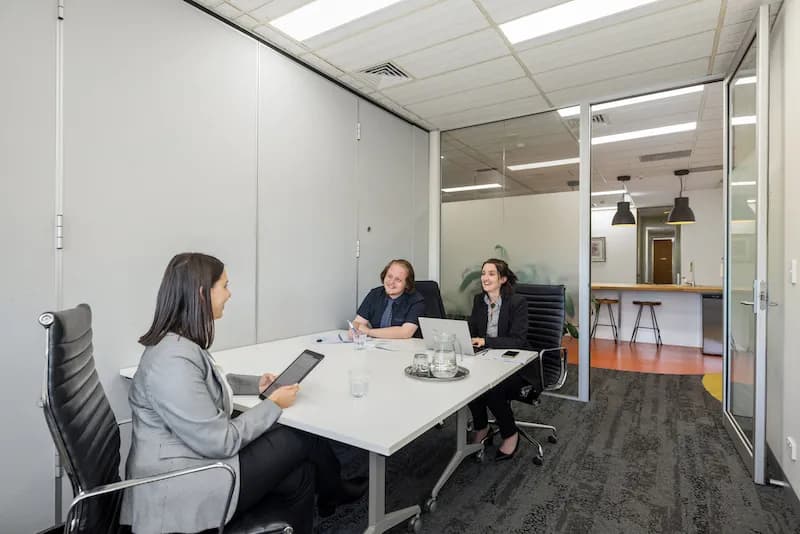 Three people sit around a white table in a modern office meeting room. Two people are on one side, facing the third person. There are laptops and notebooks on the table. Glass doors reveal a hallway and a counter with stools in the background.