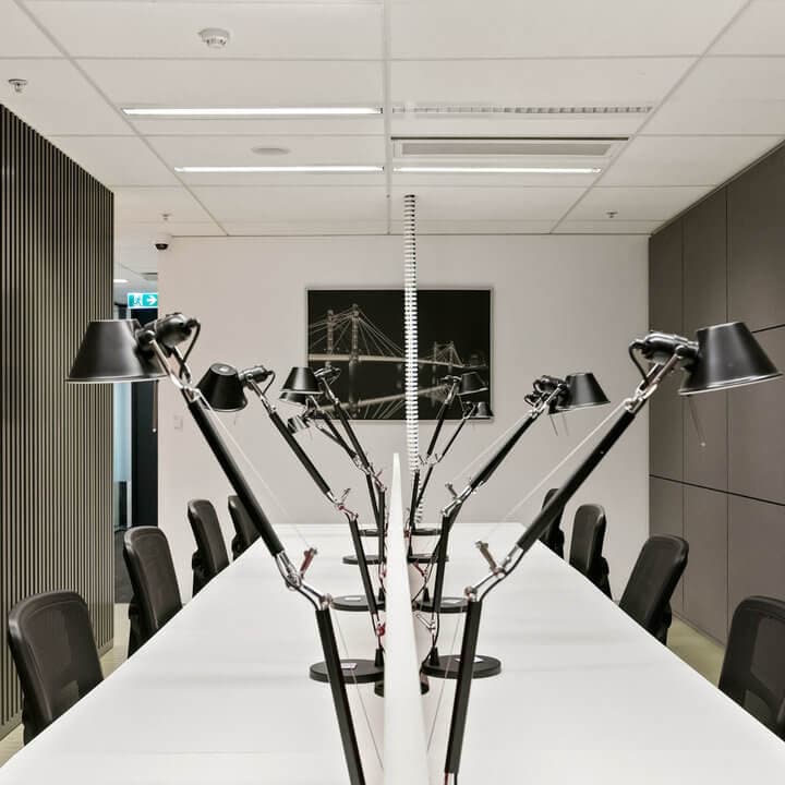 A modern conference room with a long white table, surrounded by black chairs. Multiple black desk lamps are arranged symmetrically along the table. A black-and-white photo is on the wall in the background. The room is well-lit and minimalistic.