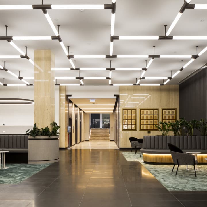 Modern office lobby with geometric lighting, wood panel walls, gray seating, and green plants. A staircase is visible in the background, and the floor is tiled in dark and green patterns.