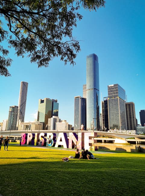 The image shows a sunny day in Brisbane, Australia. A group of people sits on a grassy area in front of large, colorful BRISBANE letters. The citys modern skyscrapers tower in the background under a clear blue sky.