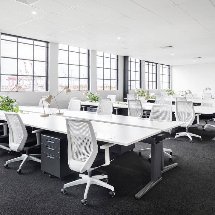 A modern office with large windows, white desks, and ergonomic white chairs. The room features desk lamps, plants, and black filing cabinets. The space is well-lit with natural light and has a minimalistic design.
