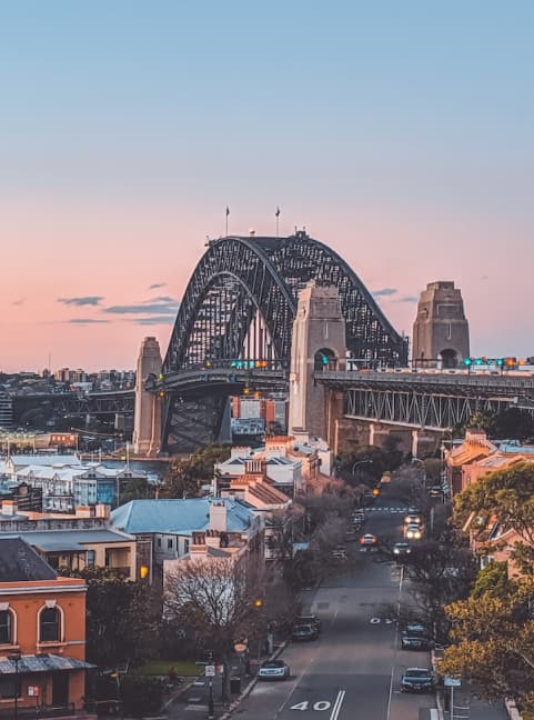View of Sydney Harbour Bridge at sunset, with a street leading towards it. The sky is pinkish, and the cityscape includes historic buildings and trees flanking the road. Traffic is light on the bridge, and street signs are visible.