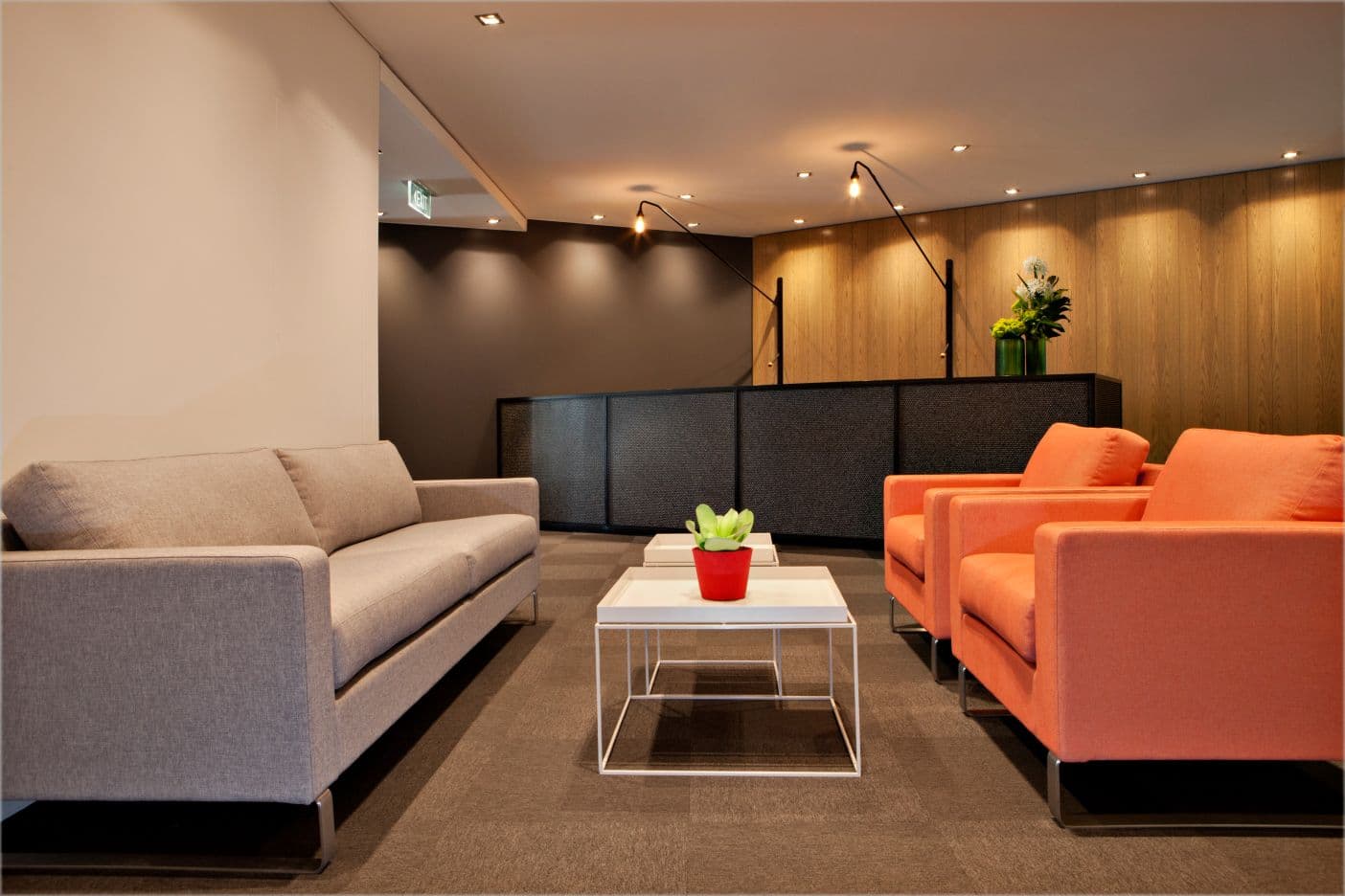 Modern waiting area with a gray sofa on the left and two orange armchairs on the right. A small white table with a potted plant in a red pot is in the center. The background features a sleek reception desk and wood-paneled wall.