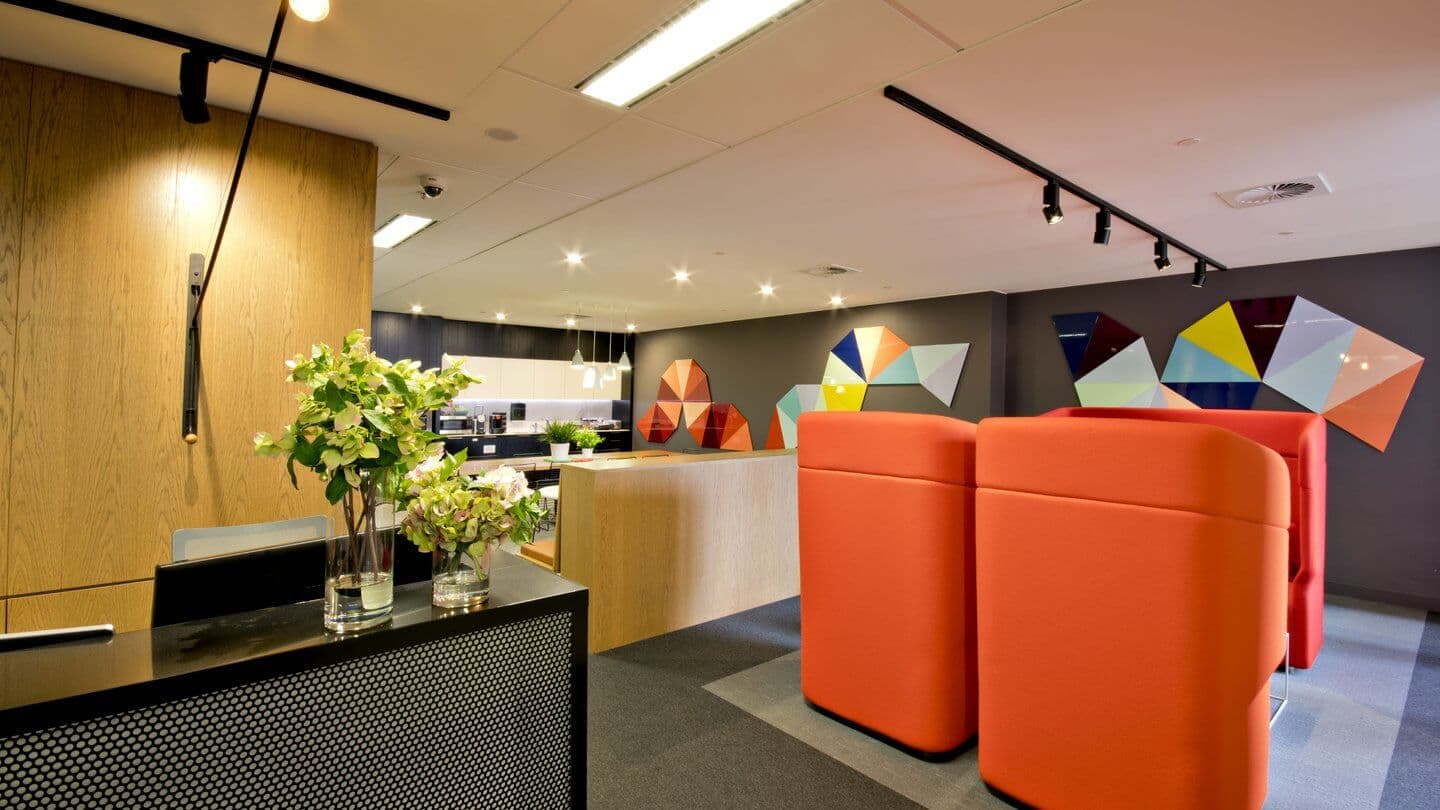 Modern office lobby with bright red seating pods, wooden reception desk, and a decorative wall with colorful geometric shapes. Overhead lighting illuminates the space, and vases with green flowers adorn the desk.